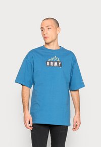 T-shirt en coton bleu à manches courtes avec un logo rectangulaire noir portant l'inscription "GRMY" et des illustrations de flammes en jaune.