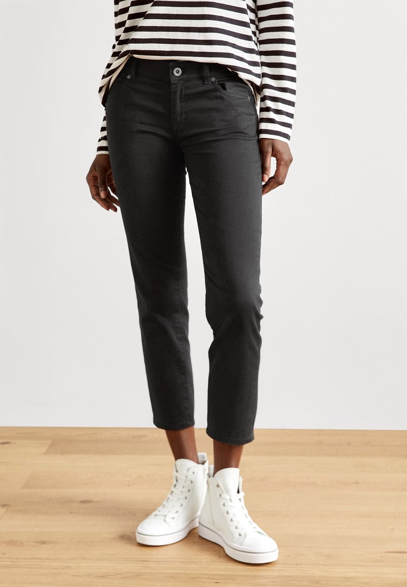 Pantalon en coton noir à coupe ajustée, longueur raccourcie, avec poches avant, porté avec des baskets blanches montantes et un t-shirt à manches longues rayé.