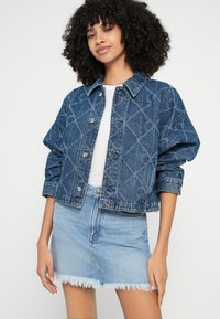 Kurtka dżinsowa z wzorem w romby, niebieska, o luźnym kroju, zapinana na guziki, z podwiniętymi rękawami, w zestawie z jasnoniebieską spódnicą dżinsową.