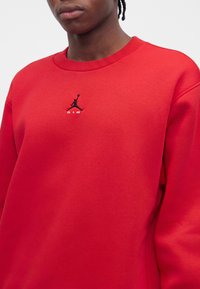 Personne portant un sweat-shirt rouge vif avec un logo Jumpman brodé en noir et le texte blanc "AIR" centré sur la poitrine.