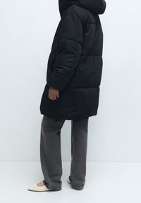 Černá péřová parka s kapucí, s dlouhým střihem, prošívanou texturou a nošená přes šedé džíny a světlé boty.