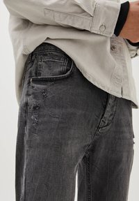 Jeans de denim negros con detalles desgastados, con un corte recto y cinco bolsillos. Combinados con una camisa de botón gris claro.