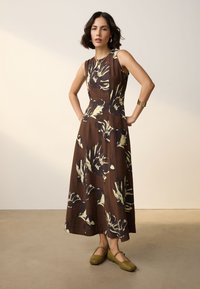 Donna in piedi con le mani sui fianchi, indossa un vestito midi marrone a fiori senza maniche e scarpe Mary Jane verde oliva su uno sfondo neutro.