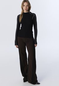 Femme debout portant un haut noir à manches longues côtelé avec des finitions blanches et un pantalon noir et marron foncé à jambes larges sur des chaussures à talons noirs.