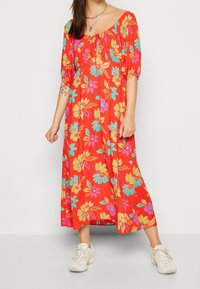 Rotes Blumenkleid im Maxiformat mit kurzen Puffärmeln, einem Rundhalsausschnitt und lebendigen Blumenmustern in Blau und Gelb. Kombiniert mit neutralen Sneakers.