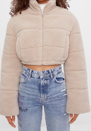 Veste courte beige en fausse peau de mouton avec fermeture éclair et manches larges, associée à un jean bleu taille haute avec texte brodé.
