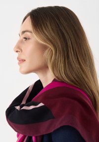 Kleurige sjaal met gedurfde patronen in tinten bordeauxrood, roze en zwart, over de schouder gedrapeerd. Zacht, gestructureerd materiaal.