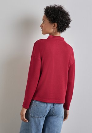 Roter gerippter Pullover mit lockerem Schnitt, hohem Kragen und langen Ärmeln. Kombiniert mit hellblauen Jeans, was einen lässigen Look zeigt.