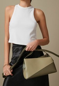 Olivgrüne Lederhandtasche mit geometrischem Design, flachem Boden und einem langen verstellbaren Trageriemen. Das Model hält die Tasche und trägt ein weißes geripptes Oberteil.