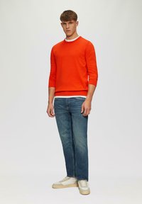 Pomarańczowy sweter z okrągłym dekoltem i długimi rękawami, noszony na białej koszuli, w komplecie z niebieskimi dżinsami i beżowymi trampkami. Prosty design, brak wzorów.