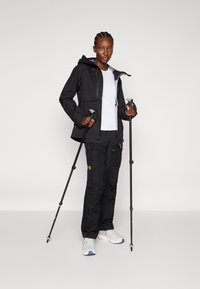 Veste noire résistante aux intempéries avec capuche, associée à un pantalon noir. La silhouette tient des bâtons de trekking, portant des chaussures blanches avec des accents bleus.