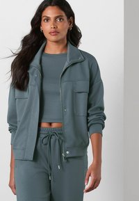 Dark green jogger set featuring a zip-up jacket with two front pockets and a cropped top, made of soft, textured material.