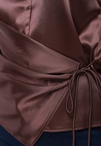 Καφέ σατέν μπλούζα με σχεδιασμό τύπου wrap και λεπτομέρεια με κορδόνι στο πλάι. Μεταξένια υφή με διακριτική γυαλάδα, συμπληρωμένη από κλείσιμο με κουμπιά.
