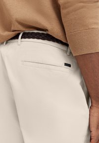 Helle beige Hosen aus strukturierter Stoffqualität, mit einer Gesäßtasche und einem kleinen schwarzen Etikett. Ein brauner geflochtener Gürtel ergänzt das Outfit.