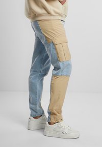 Cargo-Hosen mit einem Farbblockdesign in Hellblau und Beige, mit Seitentaschen und einem lockeren Schnitt. Getragen mit weißen Sneakers.