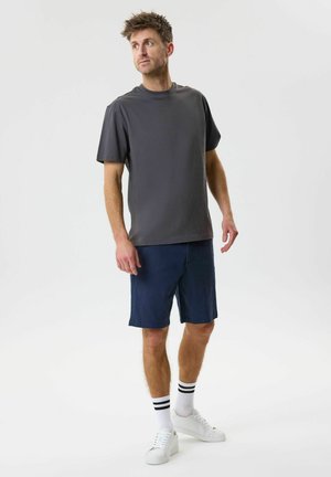 Młody mężczyzna stoi i patrzy w lewo, ubrany w ciemnoszarą koszulkę, granatowe spodenki, białe skarpetki w czarne paski i białe sportowe buty.