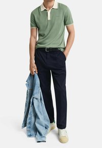 Polo vert avec un col crème, pantalon bleu marine, et une veste en denim clair tenue dans une main. Baskets blanches avec une semelle texturée.