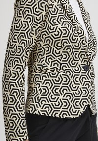 Blazer à motifs avec un design géométrique noir et crème, fermeture à un bouton, et coupe structurée. Le matériau semble lisse et tissé.