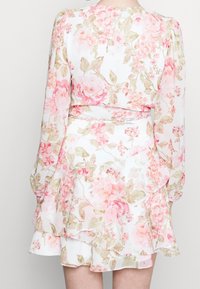 Kukkamekko, joka on valmistettu pehmeästä ja kevyestä kankaasta, jossa on vaaleanpunaisia ja vihreitä kukkakuoseja. Röyhelöhelma ja pitkät hihat viimeistelevät suunnittelun.