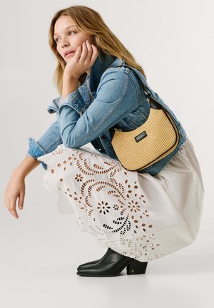 Joven agachada, vistiendo una chaqueta vaquera, falda blanca con ojales, botas negras de tacón y un bolso de hombro de paja con una etiqueta negra.