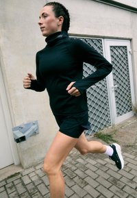 Sort atletisk langærmet skjorte med høj krave; kombineret med mørke shorts. Funktioner inkluderer en tætsiddende pasform og tekstureret stof. Løbeholdning.