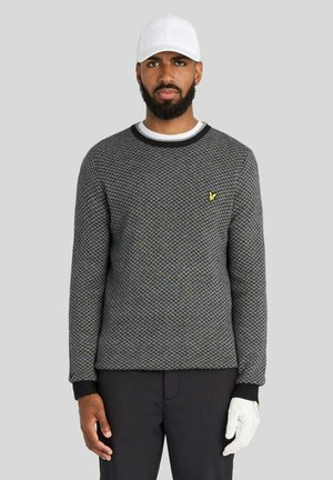 Mand iført hvid kasket, sort struktureret sweater med gul logo, sorte bukser og en hvid handske på sin højre hånd mod en grå baggrund.