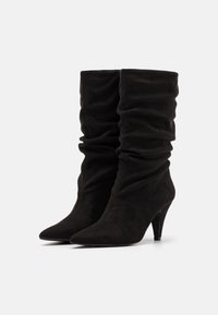 Bottes mouillées en daim noir jusqu'au milieu du mollet avec des bouts pointus et des talons aiguilles fins, présentées côte à côte sur un fond blanc.