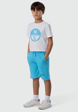 Jeune garçon debout les mains dans les poches portant un t-shirt blanc North Sails, un short bleu clair, des chaussettes blanches et des baskets blanches.
