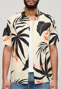 Camisa de manga corta abotonada en color crema con patrones de hojas tropicales en negro y naranja; tejido suave y corte relajado.