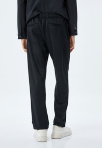 Svarta joggerbyxor med elastisk midja, sidofickor och en jämn textur. Bärs med vita sneakers mot en enfärgad bakgrund.
