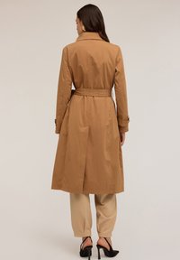 Beżowy płaszcz trench z paskiem w talii, długimi rękawami i kołnierzem. Posiada rozcięcie z tyłu, zaprezentowany na modelce w beżowych spodniach i czarnych szpilkach.