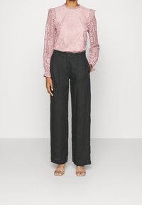 Top in pizzo a maniche lunghe di un delicato rosa, con spalle arricciate, abbinato a pantaloni neri larghi e strutturati e sandali beige chiaro.