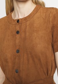 Vêtement en suède marron avec un col rond, des manches courtes et des boutons noirs sur le devant. Texture douce avec une finition lisse.