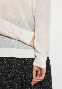 Leichter, gestrickter Pullover in Creme mit transparenter Struktur. Lange, enganliegende Ärmel und gerippte Bündchen; getragen über einem schwarzen gemusterten Rock.