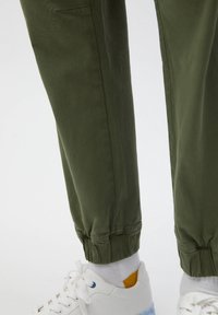 Pantalones de chándal verde oliva con puños elásticos, que presentan una textura suave y bolsillos laterales. Combinados con zapatillas blancas y calcetines de tobillo.