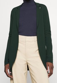 Green knit cardigan with a smooth texture, open front design, and notched collar. Worn over a dark navy turtleneck and light beige pants.