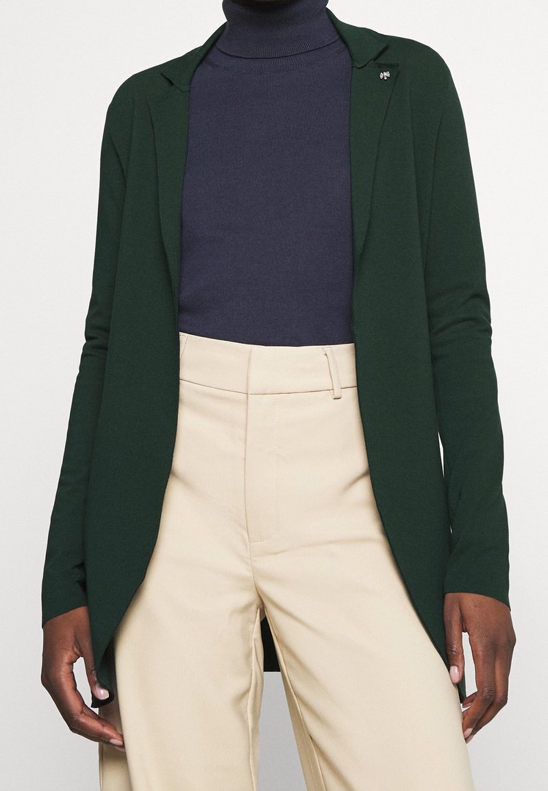 Green knit cardigan with a smooth texture, open front design, and notched collar. Worn over a dark navy turtleneck and light beige pants.