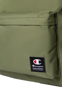 Oliwkowa torba z materiału o gładkiej fakturze, z przednią kieszenią oraz prostokątną naszywką z logo w czerwonych i czarnych akcentach.