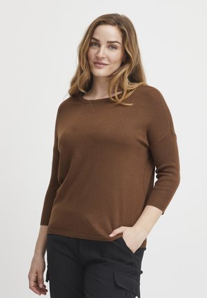 Maglione marrone a maglia con maniche a tre quarti e scollatura tonda. Texture morbida e vestibilità rilassata. Abbinato a pantaloni scuri con le mani in tasca.
