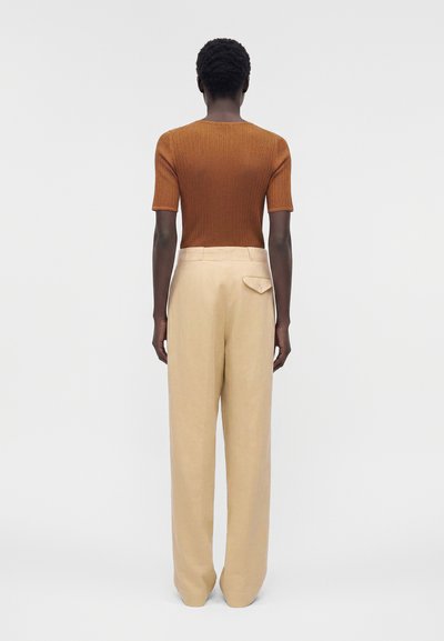 Άτομο που στέκεται με την πλάτη, φοράει καφέ ριμπ μπλουζάκι με κοντά μανίκια και μπεζ παντελόνι με υψηλή μέση, φαρδιά μπατζάκια και πίσω τσέπη με καπάκι.