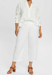 Vit linneblus med V-ringning och lång ärm, matchad med vita linnebyxor med vida ben. Bruna sandaler med klack fullbordar outfiten.