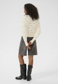 Pull en tricot crème avec un motif de losanges ouvert, associé à un short en denim gris et des bottes noires jusqu'au genou avec des détails de boucles.