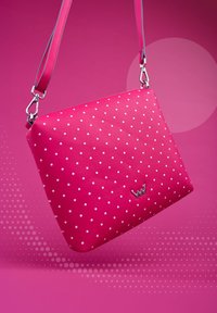 Růžová kabelka s puntíky a hladkým koženým povrchem, se lesklým stříbrným logem a nastavitelným popruhem, umístěná na živém růžovém pozadí.