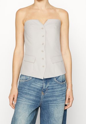 Szary, bezramiączkowy top zapinany na guziki, z dwoma przednimi kieszeniami, noszony z luźnymi niebieskimi jeansami oraz srebrną bransoletką z zawieszką w kształcie serca na prawej ręce.