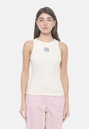 Giovane donna con lunghi capelli scuri che indossa un top bianco a coste senza maniche con un piccolo logo colorato sul petto e pantaloni rosa chiaro.