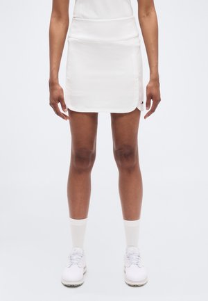 Hvid knælængde nederdel i glat, let stof med en talje. Kombineret med hvide sportssko og synlige hvide sokker.