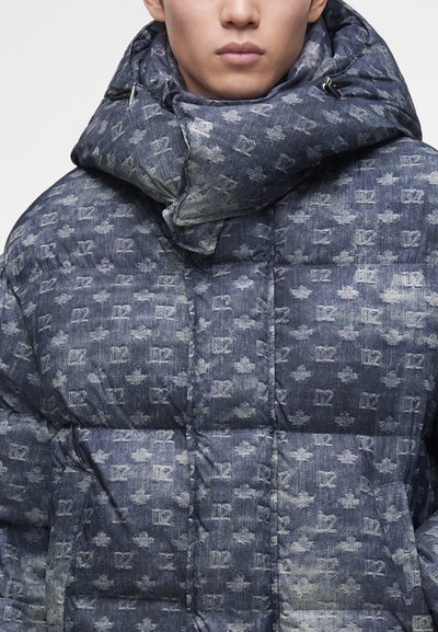 Gros plan d'une personne portant une veste matelassée bleue avec capuche, présentant un motif répétitif de "V2" et des motifs de feuilles.