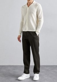 Cardigan a maniche lunghe in maglia aperta di colore crema, con colletto e bottoni, abbinato a pantaloni scuri e sneakers bianche su un pavimento grigio.