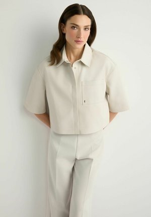 Kobieta o średniej długości brązowych włosach, ubrana w kremową koszulę z krótkim rękawem i kołnierzykiem oraz dopasowane spodnie z wysokim stanem, stojąca na tle białej ściany.