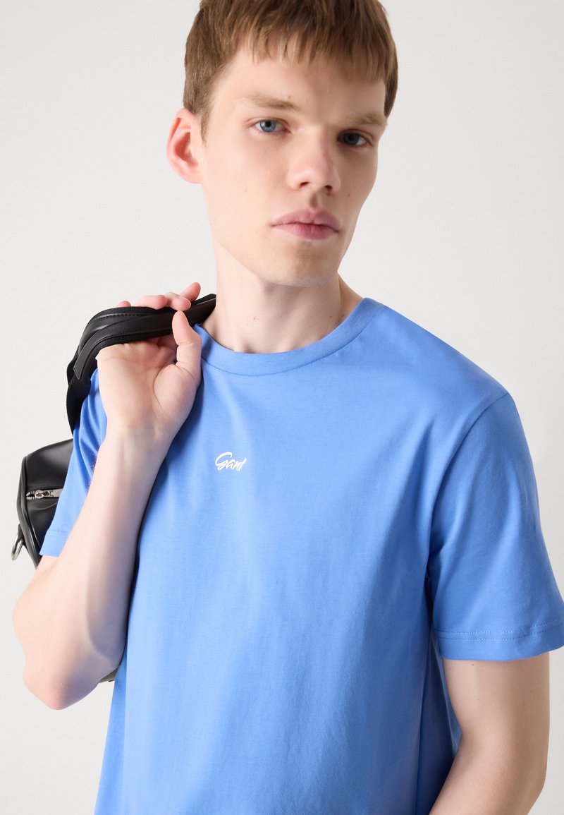 Jasnoniebieska bawełniana koszulka z okrągłym dekoltem, krótkimi rękawami i białym haftowanym logo na piersi. Model trzyma czarny pasek torby.